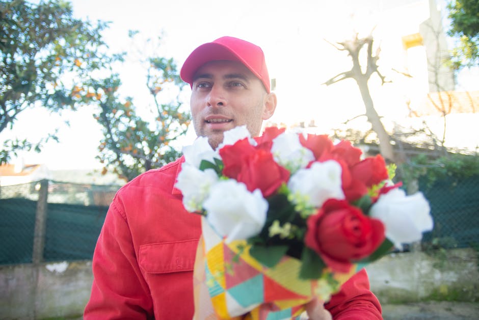 A male florist wearing a red cap and matching red uniform holds a vibrant bouquet of fresh flowers outdoors. The bouquet features red and white roses, complemented by green foliage, wrapped in colorfu