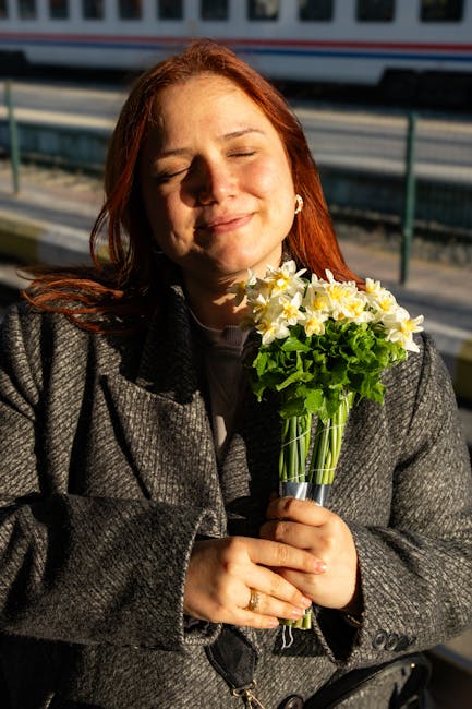 A woman with long, reddish-brown hair closed her eyes and a gentle smile on her face, holding a bouquet of fresh white and yellow daffodils with green leaves in a clear glass vase. She is dressed in a