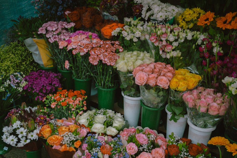 A colorful display of fresh floral arrangements arranged in white and green pots and wrapped bouquets, featuring a variety of flowers including pink roses, yellow roses, white roses, purple asters, wh