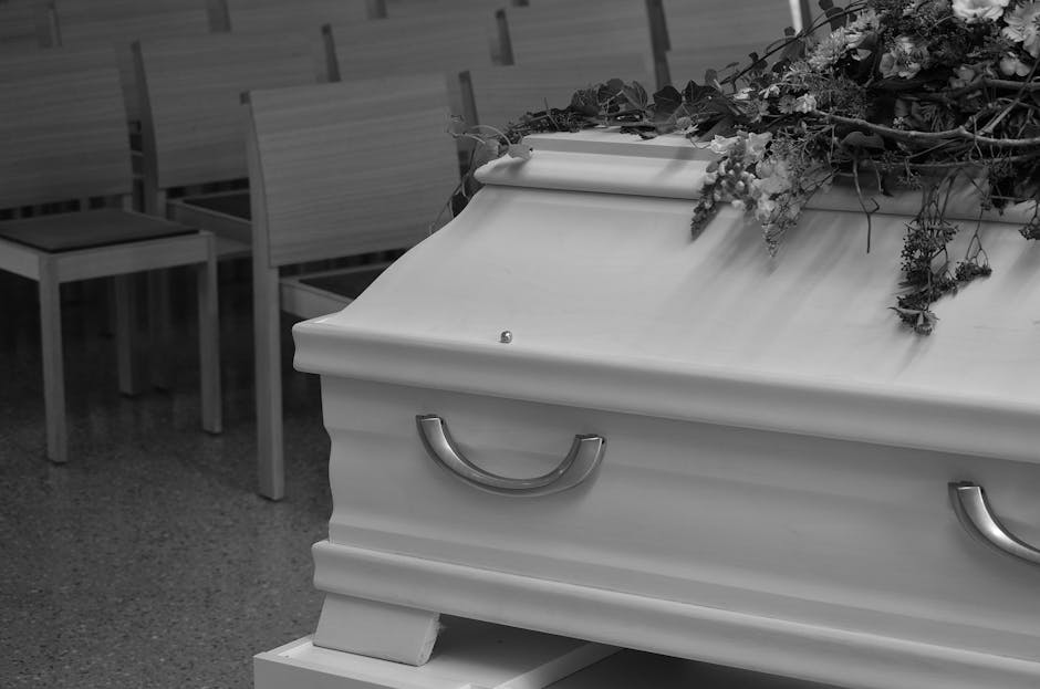 A white casket in a funeral setting, with silver handles and a decorative top. Resting on a stand, the casket is adorned with a floral arrangement featuring various flowers and greenery draped across