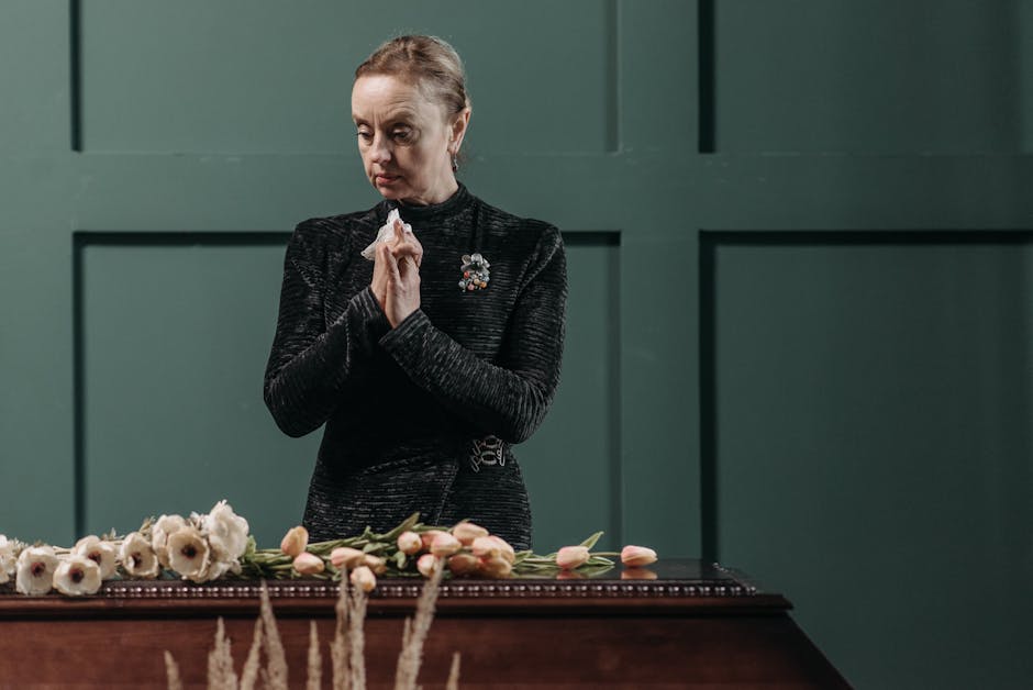 A woman in a black, textured long-sleeve dress stands solemnly behind a floral arrangement, holding a tissue with her hands pressed together in a prayer-like gesture. In front of her, there is a table