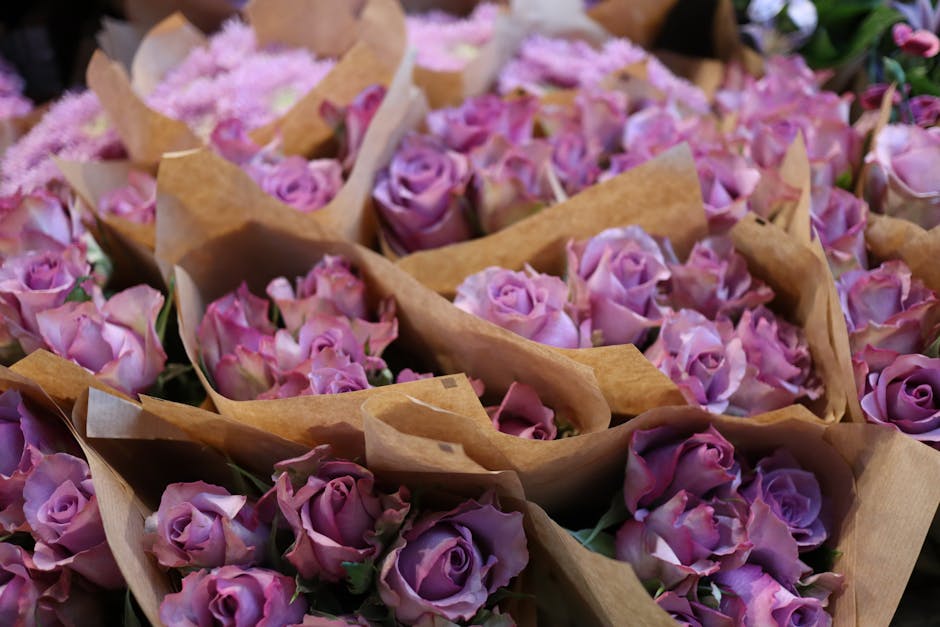 A collection of fresh pink and lavender roses arranged in small bouquets, wrapped in light brown kraft paper. The roses have tightly packed petals with a soft, velvety texture and green stems visible