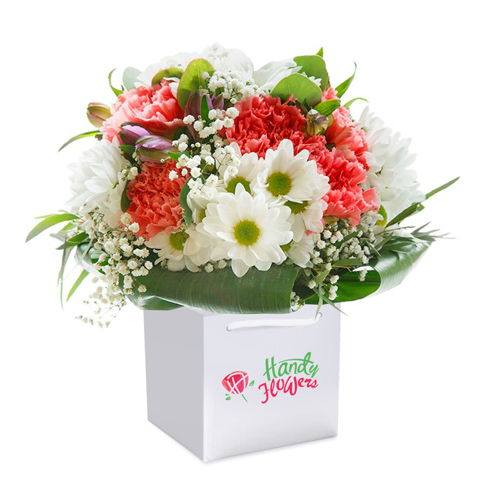 Presented upright in a spotless white gift bag, this dazzling bouquet brings together the best of Chingford's floral artistry. The arrangement centres on an exuberant gathering of peach and coral carnations, their plentiful ruffled petals creating a sumptuous texture that feels both inviting and festive. Bright white daisy-like chrysanthemums, each boasting a vivid lime-green core, add contrast and freshness-much like the sparkling mornings along Station Road. Wisps of delicate gypsophila, or baby's breath, weave a gentle, cloudlike softness throughout, while a handful of purple-tinged, still-closed buds suggest promise and depth, offsetting the arrangement's brighter hues. Framing all of this is a luxuriant collar of glossy, deep green foliage, its broad leaves artfully fanned to give the bouquet both structure and grace. The polished white bag, finished with sturdy ribbon handles, speaks of convenience and refined presentation-ideal for swift delivery across Chingford or as a hand-gifted surprise on a birthday or anniversary. Set against a brilliant pure white backdrop, this image highlights every facet of freshness and craftsmanship, reflecting the care, attention to detail, and local pride that defines Chingford's beloved independent florists.