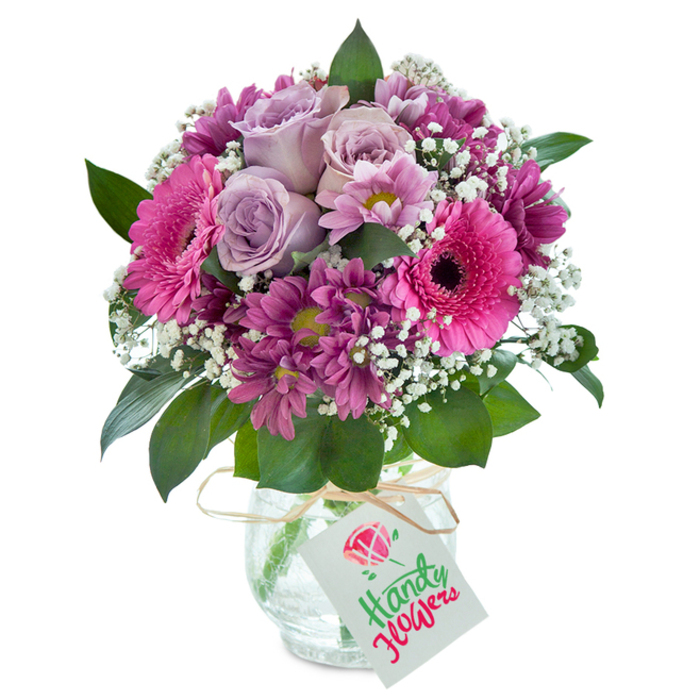 This exquisite bouquet, arranged by skilled Chingford florists, showcases a harmonious blend of pinks and purples, set gracefully against a pure white background to emphasize every nuanced tone and texture. At the heart of the bouquet, soft lavender roses bloom, their delicately curled petals suggesting the first gentle breeze of spring along Station Road. Embracing these are vibrant fuchsia Gerbera daisies-wide, open, and full of cheerful promise-paired with clusters of deep, magenta chrysanthemums whose velvety petals and subtle green centers instill layers of visual interest and depth. Interwoven sprigs of fluffy white baby's breath lend a refreshing lightness, reminiscent of the shifting skies above Connaught Water. Rich, dark green leaves arc around the arrangement, grounding the playful palette in a natural, woodland tranquility. The flowers nestle in a clear glass vase featuring a subtly crackled texture, a small design flourish that hints at the artisan care behind every local delivery. A light brown raffia ribbon wraps the vase's neck, tied with casual elegance. Suspended from the tie, a small, unadorned white gift tag hangs, inviting a hand-written note to complete the gesture. Whether gracing a home in Chingford Green, brightening an office near Highams Park, or offered as a thoughtful seasonal gift, the bouquet carries an undercurrent of warm wishes and polished artistry-an emblem of local floral excellence delivered fresh throughout the area.